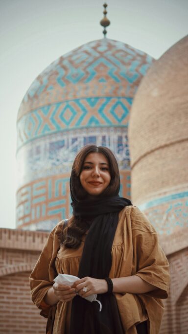 Woman in a black headscarf and loose brown clothing in front of a tiled mosque